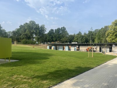 Im Bild erkennt man die grüne Liegewiese. Links im Bild befindet sich eine grüne Umkliede. Rechts im Bild steht der neue Funktionsbau mit sanitären Anlagen und Schließfächern.