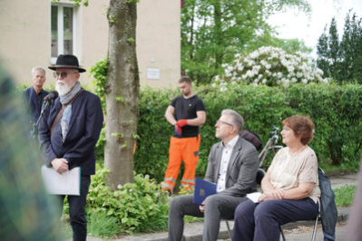 Herr Burghart und Hanna Sachs sitzen auf Stühlen und hören der Rede von Dr. Nitsche aufmerksam zu. Im Hintergrund stehen ein Musiker und ein Bauarbeiter.