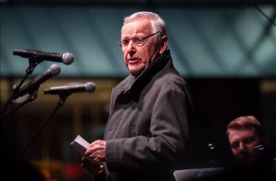 Dr. Peter Seifert, ehemaliger Oberbürgermeister der Stadt Chemnitz, steht auf einer Bühne vor einem Mikrophone. In den Händen hält er seine Rede auf A5-Papier. Er trägt einen dunkelgrauen Wollmantel und eine rahmenlose Brille. Er dreht den Kopf zur Seite und blickt ernst in die Kamera.