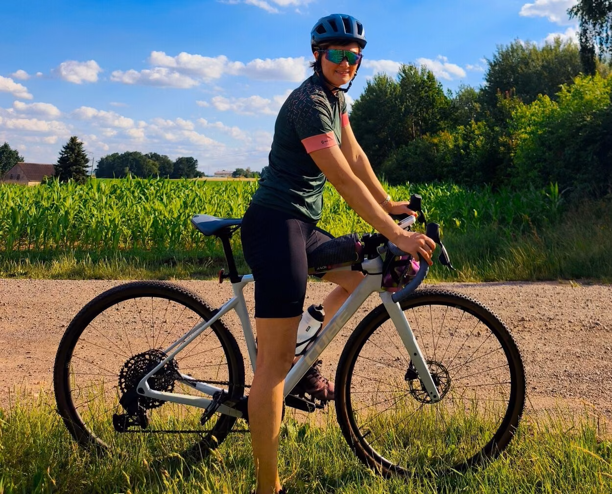 Denise Labude steht mit ihrem Fahrrad neben einem Weg an einem Felt. Sie hat eine Sonnenbrille auf und lächelt in die Kamera.