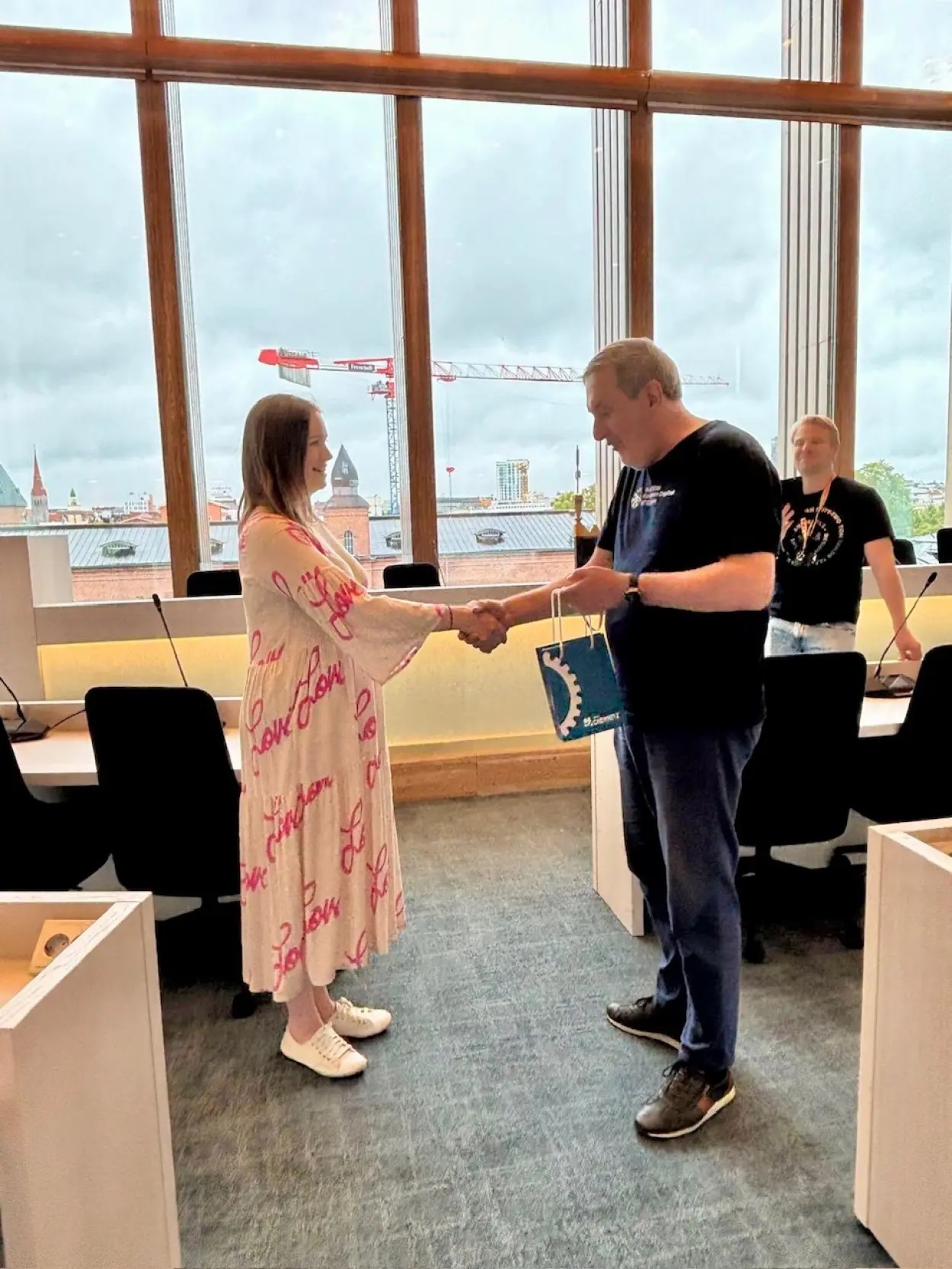 Bernd Rippert gibt sich mit der stellvertretenden B&uuml;rgermeisterin, Johanna Kirjavainen, die Hand. Bernd Rippert tr&auml;gt ein dunkles T-Shirt und eine dunkelblaue Jeans, in seiner freien Hand h&auml;lt er eine blaue Geschenkt&uuml;te der Stadt Chemnitz. Johanna Kirjavainen tr&auml;gt ein pink-rosa gemustertes, kn&ouml;chellanges Sommerkleid mit weiten &Auml;rmeln. Sie tr&auml;gt ihre Haare schulterlang in Aschblond. Im Hintergrund ist die gro&szlig;z&uuml;gige Fensterfront des Raumes erkennbar.