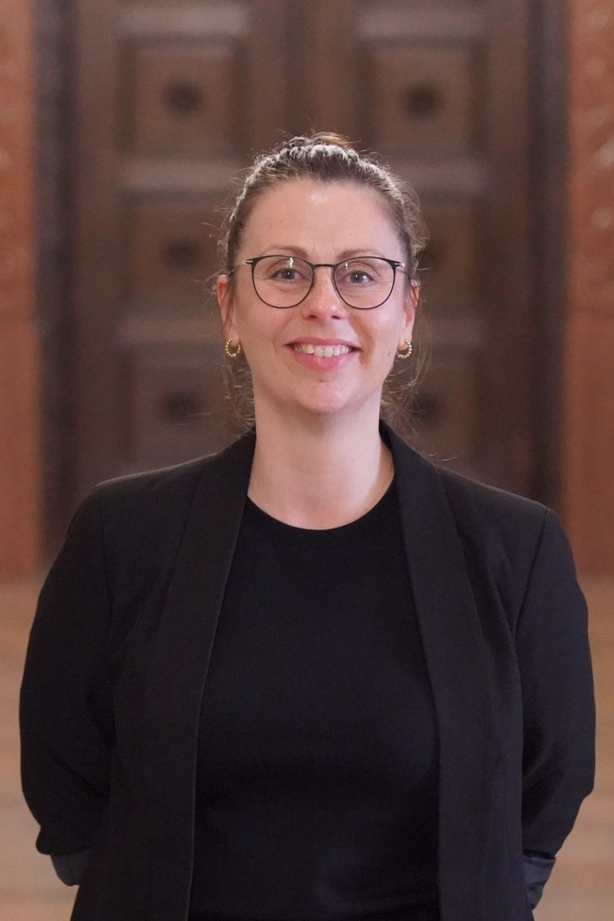 Im Bild ist eine Frau zu sehen, die in die Kamera lächelt, sie trägt eine zarte runde Brille sowie ihr braunes Haar zu einem Knoten gebunden. Bekleidet ist sie mit einem schwaruent Sakko und einem schwarzen Shirt darunter. Im Hintergund sieht man ein braune holzvertäfelte Tür, der Eingang zum Ratssal.zum