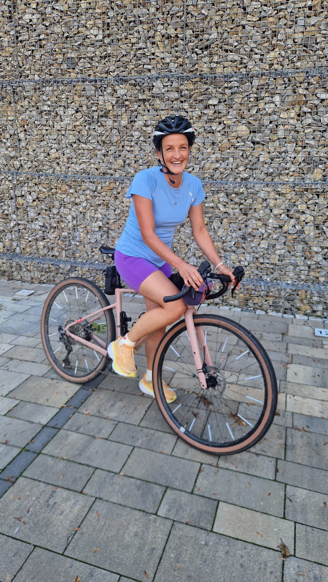 Im bild ist eine Radfahrerin zu sehen. Sie steht mit einem Bein auf dem Boden, sitzt aber im Sattel und hat die H&auml;nde am Lenkrad. Sie l&auml;chelt in die Kamera, tr&auml;gt einen Helm, ein blaues Shirt und lilafarbene Radlerhosen. Sie steht auf einem Grund aus verlegten Granitplatten, im Hintergrund ist eine Gabionen-Wand (Steine im Gitter)