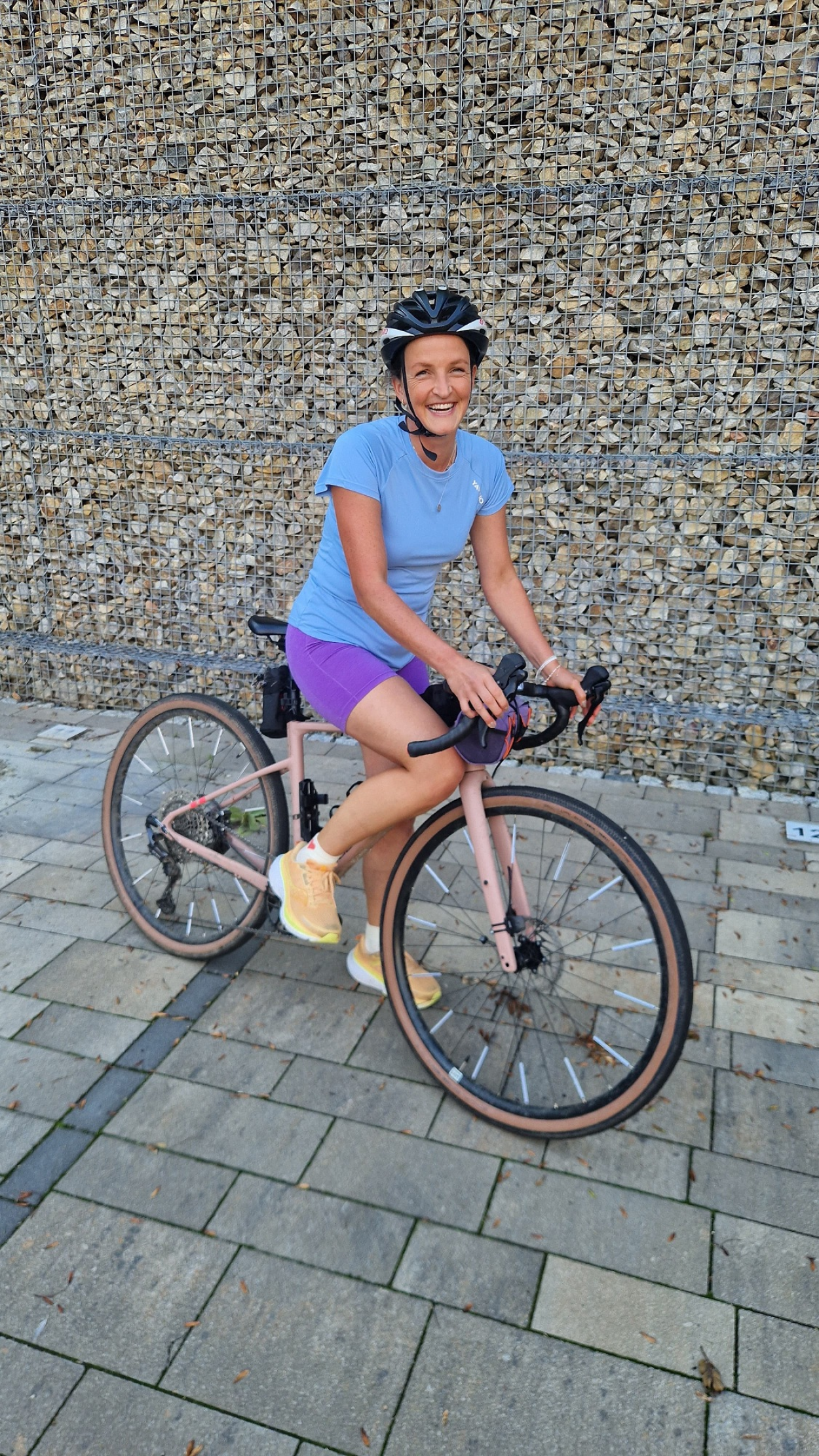 Im bild ist eine Radfahrerin zu sehen. Sie steht mit einem Bein auf dem Boden, sitzt aber im Sattel und hat die Hände am Lenkrad. Sie lächelt in die Kamera, trägt einen Helm, ein blaues Shirt und lilafarbene Radlerhosen. Sie steht auf einem Grund aus verlegten Granitplatten, im Hintergrund ist eine Gabionen-Wand (Steine im Gitter)
