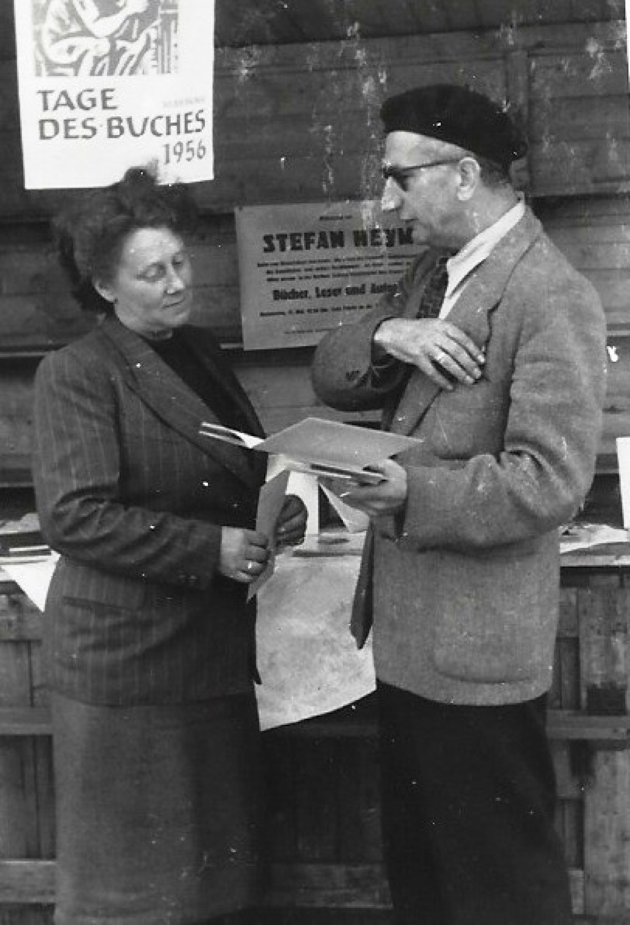 Das Ehepaar Karl und Marianne Otto ist gerade in ein Gespräch vertieft. Karl Otto hält ein schmales, weißes Buch aufgeklappt in einer Hand, mit der anderen fasst er sich gerade in die Brusttasche seines Jacketts. Er trägt eine Brille und eine schwarze Baskenmütze, eine schwarze Hose und ein weißes Hemd, dazu eine Krawatte. Marianne Otto hört ihm zu, sie scheint nachzudenken. Ihr dunkles Haar hat sie hochgesteckt. Sie trägt einen dunklen, langen Rock, ein schwarzes Oberteil und darüber einen dunklen Blazer mit Nadelstreifen. Über ihr hängt ein Plakat, auf dem „Tage des Buches 1956“ zu lesen ist. Im Schaufenster des Ladens hinter dem Ehepaar ist der Name Stefan Heym zu lesen.
